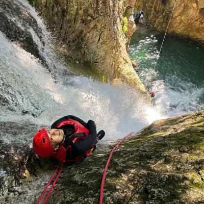 赤いヘルメットと装備を身に着けた人がロープを使って滝を登り、ゴツゴツした岩に囲まれています。背景では、もう一人の冒険者が輝く水たまりの近くで懸垂降下し、水上キャニオニングのスリルを体験しています。