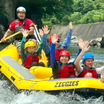 ヘルメットとライフジャケットを着た5人のグループが、黄色いゴムボートに乗って笑顔で手を挙げながら、緑と岩に囲まれた川で(水上)みなかみラフティングを楽しんでいます。