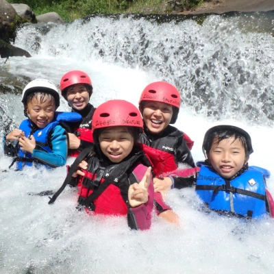 みなかみウォーターアドベンチャーを満喫しながら、激流の川で水しぶきをあげて楽しむ子供と大人のグループ。Group of kids and adults enjoying a family Minakami water adventure while splashing in a white water river.