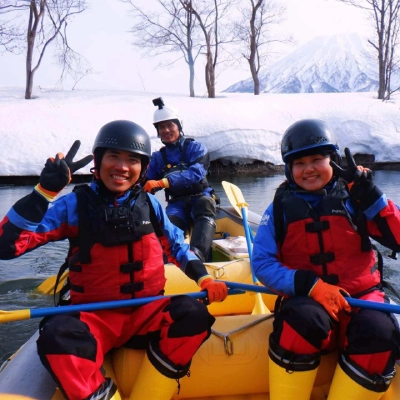 群馬県みなかみの雪景色と山を背景にした冬のラフティング体験で笑顔の参加者たち。, Winter river rafting tour in Minakami, Japan, featuring smiling participants posing in a raft surrounded by snowy landscapes and a scenic mountain backdrop, enjoying a cold-weather outdoor adventure.